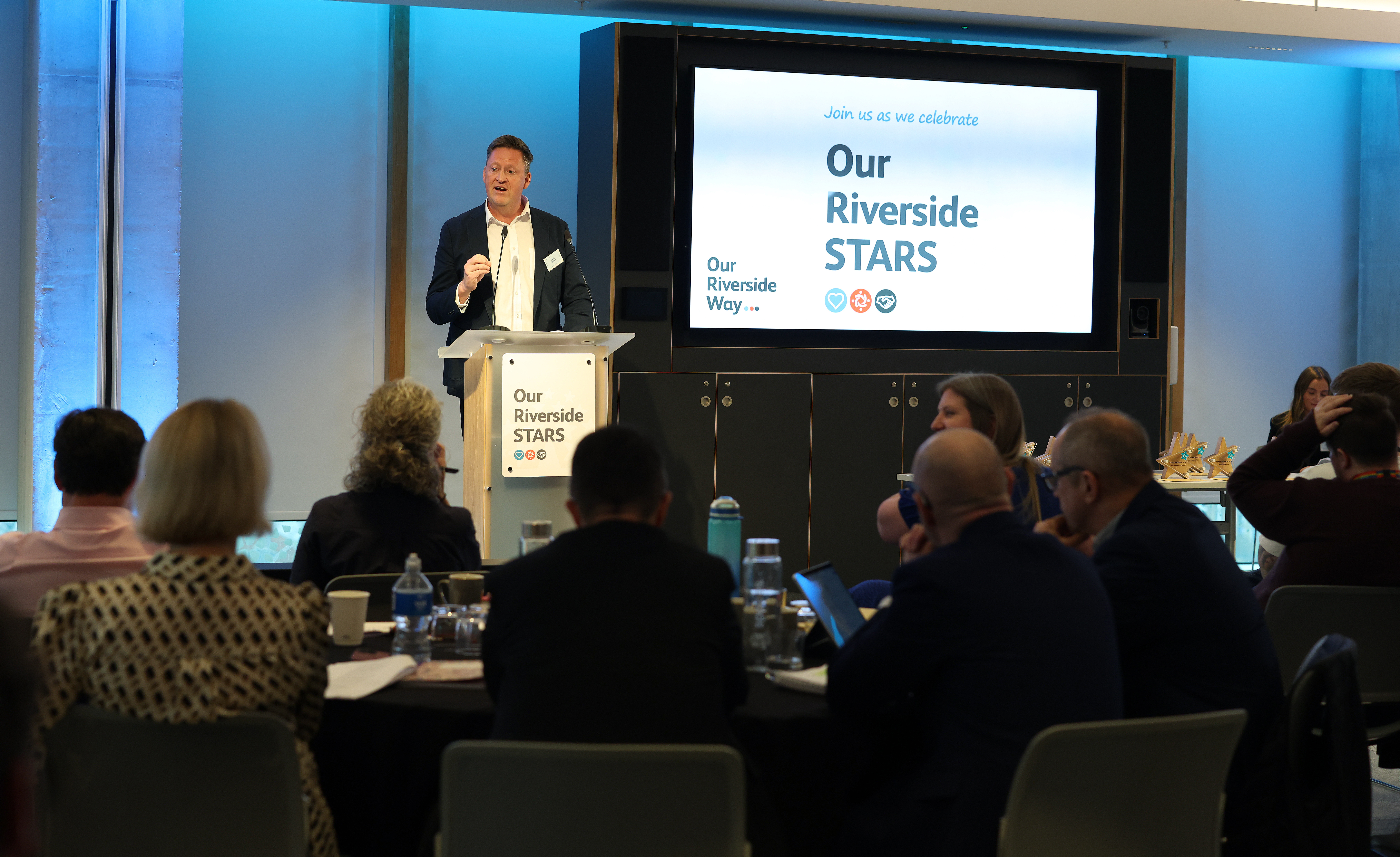 A man stands behind a podium in front of a screen which reads 'Our Riverside STARS'