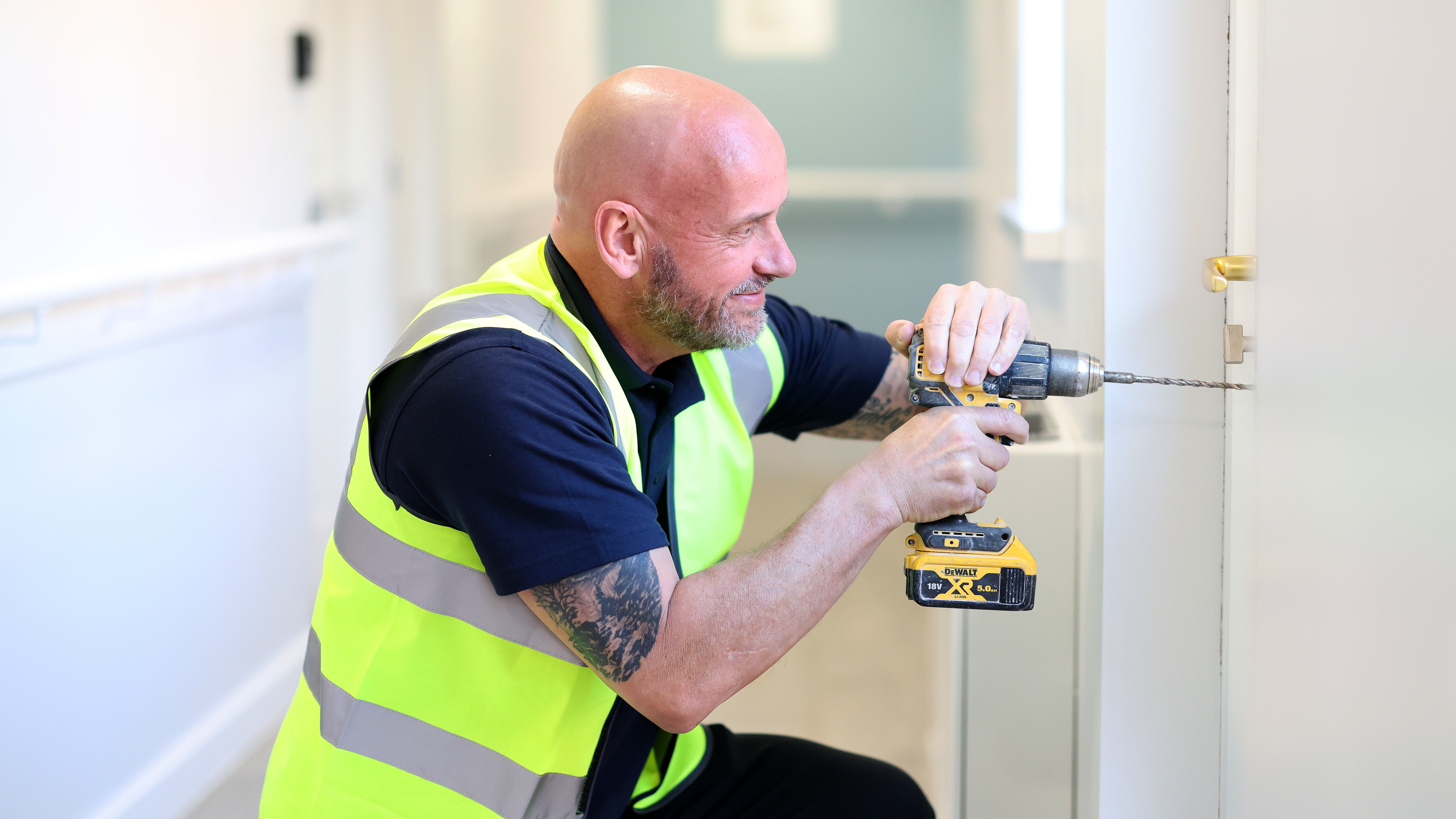 A Riverside repairs technician wearing a high-visibility vest using a power drill to fix a door hinge inside a building